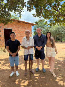 Equipe de quatro pessoas posando com idoso em frente a casa de tijolos em área rural para produção audiovisual