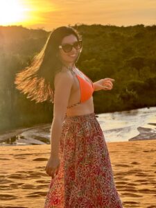 Mulher sorridente de biquíni laranja e saia estampada posando em praia ao pôr do sol dourado com vegetação ao fundo