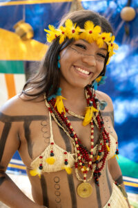 Mulher jovem com traje tradicional indígena, cocar amarelo, pintura corporal e colares coloridos sorrindo